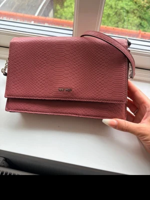 Rosa axelväska från Nine West - Säljer en snygg rosa axelväska från Nine West med strukturmönster som liknar ormskinn. Väskan har ett stilrent, rektangulärt format lock och en axelrem med kedjedetaljer. Perfekt för dig som vill ha en trendig och praktisk väska till vardagen.