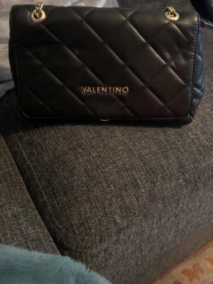 Svart quiltad axelväska Valentino - Snygg svart axelväska från Valentino med quiltat mönster och guldfärgade kedjor som axelrem. Väskan har en stilren rektangulär form och logga i guld framtill. Perfekt för dig som vill ha en trendig och elegant accessoar.