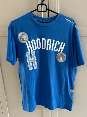 Blå HOODRICH t-shirt med tryck - Snygg blå t-shirt från HOODRICH med vita och grå tryckdetaljer framtill samt logga på axeln. T-shirten har klassisk passform, rund halsringning och är tillverkad i mjuk bomull. Perfekt för en streetwear-look.