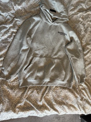 Grå Essentials Fear of God hoodie - Snygg grå hoodie från Essentials Fear of God med svart logga på bröstet. Klassisk passform med stor känguruficka och huva. Perfekt för en avslappnad streetwear-look. Mjuk bomullsblandning som känns skön mot huden.