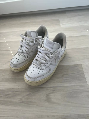 Vita Nike Air Force 1 sneakers - Klassiska vita Nike Air Force 1 sneakers med lågt skaft och perforerad tåbox. Skorna har vit ovandel i läder, vit sula med gulnad kant och tydliga swoosh-loggor på sidorna. Tjocka vita skosnören och vadderad plös för extra komfort.