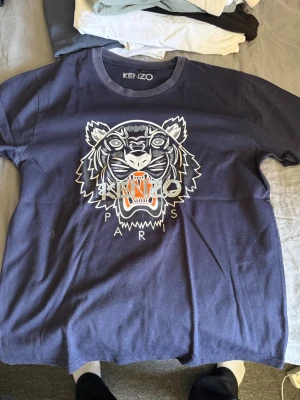 Mörkblå Kenzo t-shirt med tigertryck - Snygg mörkblå t-shirt Kenzo med det ikoniska tigertrycket och texten 'KENZO PARIS' på bröstet. Klassisk rund halsringning och korta ärmar. Perfekt för dig som vill sticka ut med en trendig och ungdomlig stil.