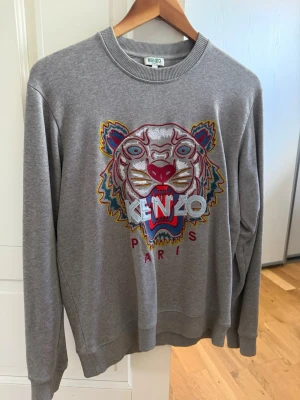 Grå Kenzo sweatshirt med tigertryck - Säljer en grå sweatshirt från Kenzo Paris med det ikoniska broderade tigerhuvudet i flera färger på bröstet. Perfekt statement-plagg för dig som gillar streetwear. Storleken är S