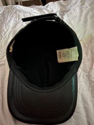 Svart keps från Gucci med GG-mönster - Snygg svart keps från Gucci med klassiskt GG-monogram i grått över hela kepsen. Skärmen är i svart läder och baksidan har justerbart band. Perfekt accessoar för en trendig och exklusiv look.