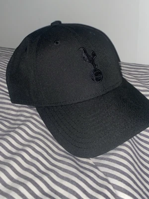  Tottenham keps  - Oanvänd Snygg svart keps från New Era med Tottenham Hotspur-logga broderad framtill i samma färg. Klassisk 9FORTY-modell med böjd skärm och justerbar rem bak. Tillverkad i bomull för skön passform och stilren look.