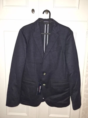 Mörkblå kavaj med två knappar från Tommy Hilfiger - Stilren mörkblå kavaj med två knappar framtill och tre fickor. Klassisk passform med smala slag och diskret struktur i tyget. Perfekt för dig som gillar en enkel men snygg look. Nypris 5500kr.