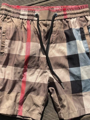 Rutiga badbyxor från Burberry - Snygga badbyxor från Burberry med klassiskt rutmönster i beige, svart, vitt, rött och blått. Modellen har resår i midjan med svart dragsko och sidofickor. Perfekta för stranden eller poolen och tillverkade i snabbtorkande material.