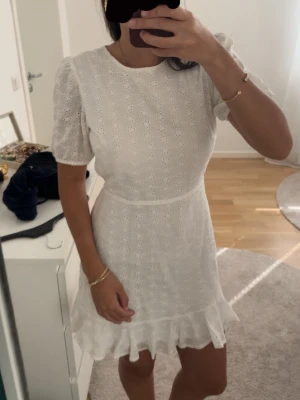 Vit kort klänning med volang - Söt vit kort klänning i bomull med broderade blommor, puffärm och volangkant nedtill. Klänningen har rund halsringning och markerad midja för en feminin siluett. Perfekt för sommarens alla tillfällen.