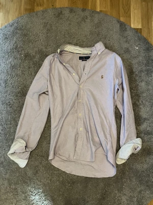 Ljuslila skjorta från Ralph Lauren - Klassisk ljuslila skjorta från Ralph Lauren med button-down krage och broderad logga på bröstet. Skjortan har långa ärmar, randigt tyg på insidan av kragen och knappar framtill. Perfekt för en clean och stilren look.