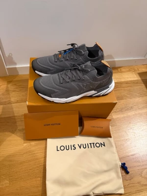 Grå Louis Vuitton sneakers - Snygga grå sneakers från Louis Vuitton med vit och svart sula. Skorna har en modern design med meshdetaljer och LV-logga på sidan. Orange detalj på hälen och blå detalj vid snörningen. Perfekta för dig som vill ha något exklusivt och trendigt.