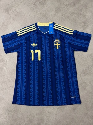 Blå Sverige fotbollströja Adidas - Säljer en blå Sverige fotbollströja från Adidas med gula detaljer och nummer 17. Tröjan har trycket 'GYÖKERES' på ryggen, tre gula ränder på axlarna och ett mönster i olika blå nyanser. Tillverkad i lätt och ventilerande material, perfekt för match eller träning.