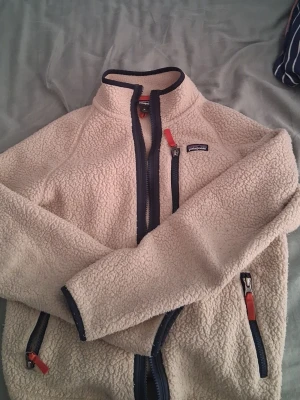 Beige fleecejacka från Patagonia - Mysig beige fleecejacka från Patagonia med marinblå dragkedjor och röda detaljer. Jackan har hög krage, två sidofickor och en bröstficka med dragkedja. Perfekt för kalla dagar och outdoor-häng.