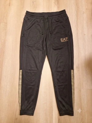 EA7 Emporio Armani Mjukisbyxor / Joggers (Svart/Grå) – Stl L ⚡🖤 - Säljer ett par riktigt snygga och bekväma mjukisbyxor/joggers från EA7 Emporio Armani! Byxorna är stilrent svarta med diskreta grå/silvriga logoband med "Emporio Armani"-text längs sidorna samt EA7-logga på låret. De har elastisk midja med snörning, praktiska sidofickor med dragkedja och en bakficka. Perfekta för en sportig street-look, gymmet eller till vardags. Märke: EA7 Emporio Armani Storlek: L (Large) Färg: Svart med gråa/silvriga detaljer Skick: Mycket gott begagnat skick!