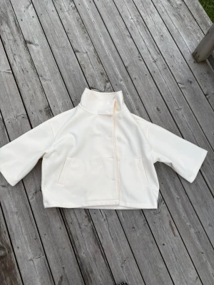 Vit kort kappa med hög krage - Stilren vit kappa i kort modell med hög krage och trekvartsärmar. Kappan har dold knäppning framtill och två fickor. Materialet ser ut att vara mjukt och något tjockare, perfekt för kyligare dagar. Enkel och modern design som passar till många outfits.