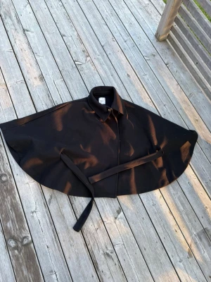 Svart cape med bälte - Stilren svart cape med klassisk krage och knäppning framtill. Modellen har ett tillhörande bälte i midjan för en snygg siluett.