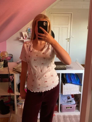 Vit blommig topp från Kappahl - Söt vit topp från Kappahl med små rosa blommor och ribbad struktur. Toppen har korta ärmar, vågig kant vid ärmslut och nederkant samt rundad halsringning. Perfekt för en somrig och fräsch look.
