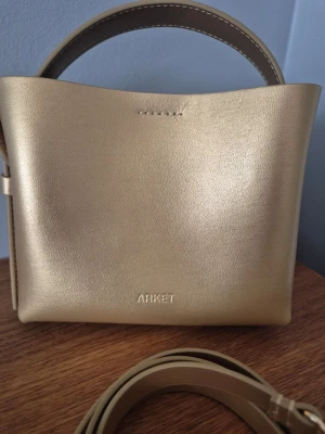 Guldig handväska från ARKET - Snygg guldig handväska från ARKET i slätt läder. Väskan har en stilren, boxig form med ett kort handtag och en avtagbar axelrem. Perfekt för dig som vill ha en trendig accessoar med minimalistisk design.