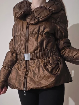 Tuzzi brun glansig pufferjacka 🤎 höstjacka med puffkrage - En otroligt fin och stilren jacka från Tuzzi med glansig finish som ger ett lyxigt och elegant uttryck ✨ Jackan har en stor mysig puffkrage, dragkedja framtill och en dekorativ spännedetalj som framhäver silhuetten på ett feminint sätt 🤎 Perfekt som höstjacka för dig som vill ha en varm, trendig och samtidigt uppklädd look 🍂  📏 Mått ca (ej utsträckt): Axelbredd: 41 cm Bröstvidd (armhåla till armhåla): 42,5 cm Längd: 63 cm 💌 Skriv gärna vid frågor eller om fler bilder önskas. Bundle-rabatt finns💕