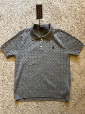 Grå kabelstickad polotopp från Polo Ralph Lauren - Snygg grå kabelstickad polotopp från Polo Ralph Lauren med korta ärmar och klassisk krage. Toppen har två knappar framtill och den ikoniska loggan broderad på bröstet. Perfekt för en clean och stilren look.