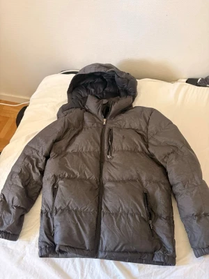 Grå pufferjacka från Polo Ralph Lauren - Säljer en grå pufferjacka från Polo Ralph Lauren med huva och tre dragkedjeförsedda fickor. Jackan har diskret logga på bröstet och breda kanaler för extra värme. Perfekt för kalla vinterdagar! 