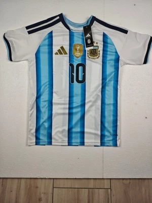 Argentina Messi matchtröja nr 10 - Snygg Argentina matchtröja från Adidas med Messi och nummer 10 på ryggen. Tröjan har vita och ljusblå vertikala ränder, svarta detaljer på axlarna och officiella klubbmärken på bröstet. Tillverkad i lätt och ventilerande material, perfekt för fotboll eller träning.