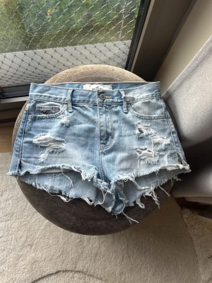 Ljusa jeansshorts från Hollister - Säljer ett par ljusblå jeansshorts från Hollister med råa, fransiga kanter och flera slitningar framtill och baktill. Klassisk femficksmodell med knappgylf och broderade detaljer på bakfickorna. Perfekta för en avslappnad och trendig look.
