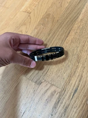 Svart armband med metallinslag - Snyggt svart armband med detaljer i silverfärgad metall. Armbandet har en modern design med dubbla svarta band och metallinslag i mitten. Stängs med ett robust metallspänne. Perfekt accessoar för en stilren look.