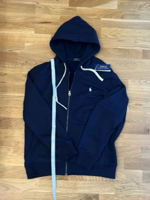Mörkblå hoodie från Polo Ralph Lauren - Snygg mörkblå hoodie med dragkedja från Polo Ralph Lauren. Klassisk design med vit dragsko i huvan, ribbade muddar och fickor framtill. Liten broderad logga på bröstet. Perfekt för en avslappnad och stilren look.