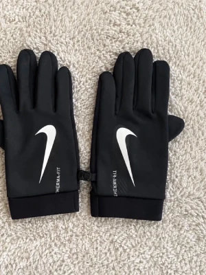 Svarta Nike Therma-Fit handskar - Snygga svarta handskar från Nike med vitt swoosh-tryck och texten THERMA-FIT på ovansidan. Handskarna har en slimmad passform och muddar vid handleden. Perfekta för kyliga dagar när du vill hålla händerna varma och samtidigt se sportig ut.
