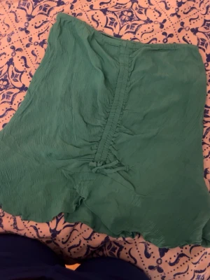 Grön omlott blus med knyt - Snygg grön blus i lätt och skrynkligt tyg med omlott och knyt framtill. Perfekt för dig som gillar en avslappnad men trendig stil. Blusen har korta ärmar och en luftig känsla, vilket gör den skön att bära under varmare dagar. (kan användas som kjol)