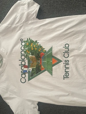 Vit Casablanca Tennis Club t-shirt - Säljer en vit t-shirt med tryck från Casablanca Tennis Club. Framsidan har ett färgglatt motiv med tennisbana, blommor och texten 'Casablanca Tennis Club'. Klassisk passform och rund hals. Perfekt för dig som gillar sportig och trendig stil. Passar även XL 