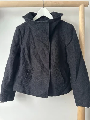 Svart kort jacka med bred krage - Stilren svart jacka med bred krage och en knappdetalj framtill. Jackan är kortare i modellen och har två fickor på sidorna. Insidan har ett mönstrat foder i vitt, svart och lila. 