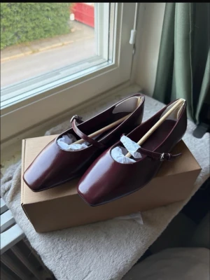 Vinröda ballerinaskor med rem - Stilrena ballerinaskor i vinrött med fyrkantig tå och klassisk rem över foten. Skorna har en elegant, minimalistisk design och är tillverkade i ett glansigt material som liknar läder. Perfekta för dig som gillar en chic och trendig look. Nypris 499kr. Ena skon är provad