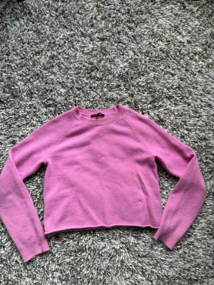 Rosa cropped kashmir stickad tröja från Maje - Snygg rosa stickad tröja med croppad passform och långa ärmar. Tröjan har ribbade muddar vid ärmslut och nederkant samt en liten broderad detalj fram och bak i vitt. 