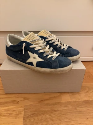 Golden Goose Superstars - Snygga blå sneakers från Golden Goose med vit stjärndetalj på sidan, vita skosnören med text och silverfärgade detaljer vid hälen. Ovandelen är i mocka och insidan är fodrad i vitt. Klassisk låg modell med slitna detaljer för en cool look.
