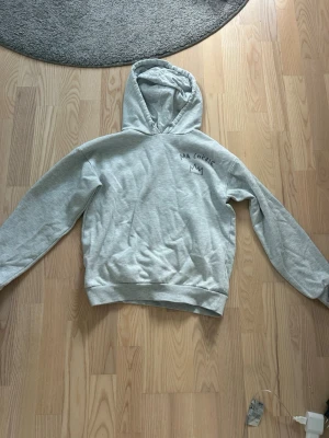 Grå hoodie med tryck 'MA CHÉRIE' - Säljer en ljusgrå hoodie med huva och trycket 'MA CHÉRIE' samt en krona på både bröstet och ryggen. Hoodien har ribbade muddar vid ärmslut och nederkant. Perfekt för en chill och avslappnad stil.