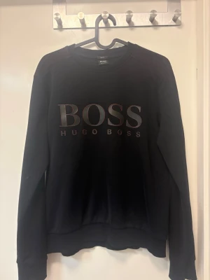 Svart sweatshirt från Hugo Boss - Svart sweatshirt med stor BOSS-logga och texten HUGO BOSS på bröstet. Tröjan har rund halsringning, långa ärmar och ribbade muddar. Perfekt för en clean och stilren look. Med röda detaljer. (Inget solblekt)