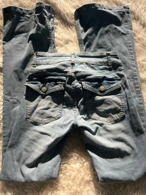 Ljusblå jeans med ficklock - Säljer ett par ljusblå jeans med raka ben och snygga ficklock bak med dekorativa knappar. Jeansen har klassisk femficksmodell och bälteshällor. Perfekta för en avslappnad och trendig look.
