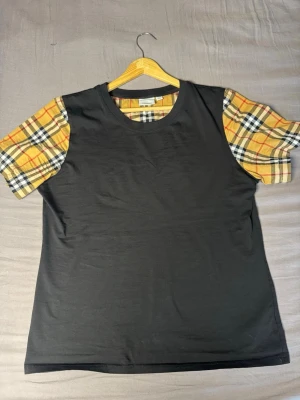 Burberry T-shirt  - Cool svart t-shirt med kortärmade ärmar i gult och svart rutigt mönster. T-shirten har en klassisk rund halsringning och är tillverkad i mjukt material, perfekt för en avslappnad stil.