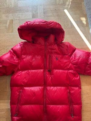 Ralph Lauren Dunjacka - Snygg röd dunjacka med huva och dragkedja framtill. Jackan har två sidofickor med dragkedja och en bröstficka. Puffig modell som håller dig varm under kalla dagar. Liten logga på bröstet i mörkblått. Perfekt för vintern!