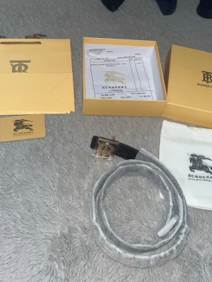 Burberry bälte med rutigt mönster - Snyggt Burberry bälte med klassiskt beige, svart och vitt rutigt mönster. Bältet har ett elegant metallspänne med Burberrys logotyp och kommer med originalpåse och box. Perfekt accessoar för att lyfta din outfit.