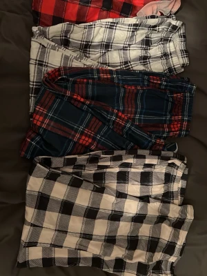 Fyra rutiga pyjamasbyxor - Säljer ett set med fyra pyjamasbyxor i olika rutiga mönster. Färgerna är röd-svart, vit-svart, blå-röd och vit-svart. Byxorna har avslappnad passform och resår i midjan för extra komfort. Perfekta för mysiga hemmakvällar eller lata morgnar.