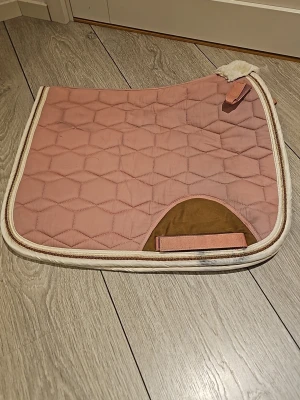 Rosa schabrak från Trinity - Säljer ett rosa schabrak från Trinity med sexkantigt quiltat mönster och beige/vita detaljer. Schabraket har en vit kant och en brun förstärkning vid sadelgjorden. Perfekt för dig som gillar stilrena hästtillbehör. Storlek Cob, andvänd ett par gånger men är i fint skick.