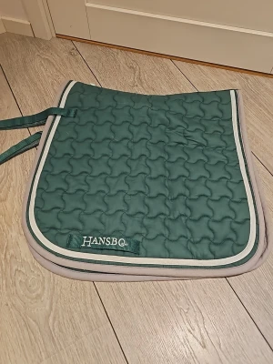 Grönt schabrak från Hansbo - Säljer ett grönt schabrak från Hansbo med vit kant och quiltat mönster. Sadelpaden har praktiska remmar för fastsättning och är tillverkad i mjukt, vadderat material. Perfekt för ridning och ger extra komfort till hästen. Storlek ponny och bara andvänd 1 gång. Dressyr schabrak