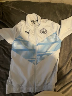 Manchester City vindjacka från Puma - Ljusblå och vit vindjacka från Puma med Manchester City-logga på bröstet. Jackan har dragkedja framtill, ståkrage och elastiska muddar vid ärmsluten. Snygga blåa och vita ränder över bröstet ger en sportig look. Perfekt för dig som gillar fotboll och streetwear.