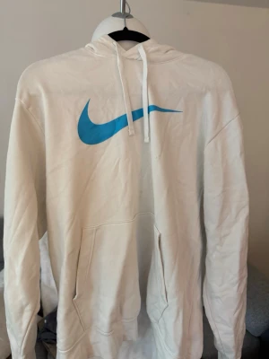 Vit hoodie från Nike med blå logga - Säljer en vit hoodie från Nike med stor blå Swoosh-logga på bröstet. Hoodien har huva med dragsko och en klassisk känguruficka framtill. Tillverkad i mjukt material som passar perfekt för en avslappnad stil.