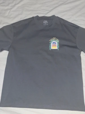 Grå t-shirt från Casablanca - Snygg grå t-shirt från Casablanca med ett färgglatt tryck på bröstet som föreställer en byggnad och gröna blad. T-shirten är gjord i 100% kammad bomull och har en klassisk rund hals. Perfekt för dig som gillar stilrena plagg med unika detaljer.