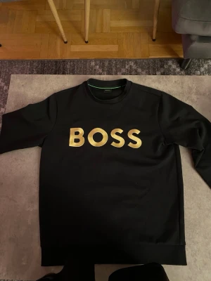 Svart sweatshirt från BOSS - Snygg svart sweatshirt från BOSS med stor guldfärgad logga på bröstet. Tröjan har rund halsringning och långa ärmar. Perfekt för dig som vill ha en enkel men ändå statement look. Materialet känns mjukt och bekvämt.