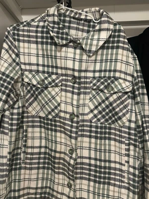 Grönrutiga overshirt med fickor - Overshirt i grön, svart och vit rutigt mönster. Två stora bröstfickor med knappar och klassisk krage. Stängs med knappar framtill och har långa ärmar. Materialet ser ut att vara mjukt och lite tjockare, perfekt för lager på lager.