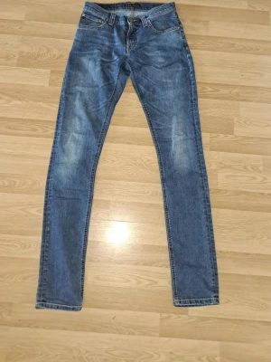 Blå slim fit jeans med slitningar -  Säljer ett par snygga Nudie Jeans i blå denim. Jeans med en klassisk passform och snyggt naturligt sliten/faded look. Bra skick   Storlek: W27 / L34 